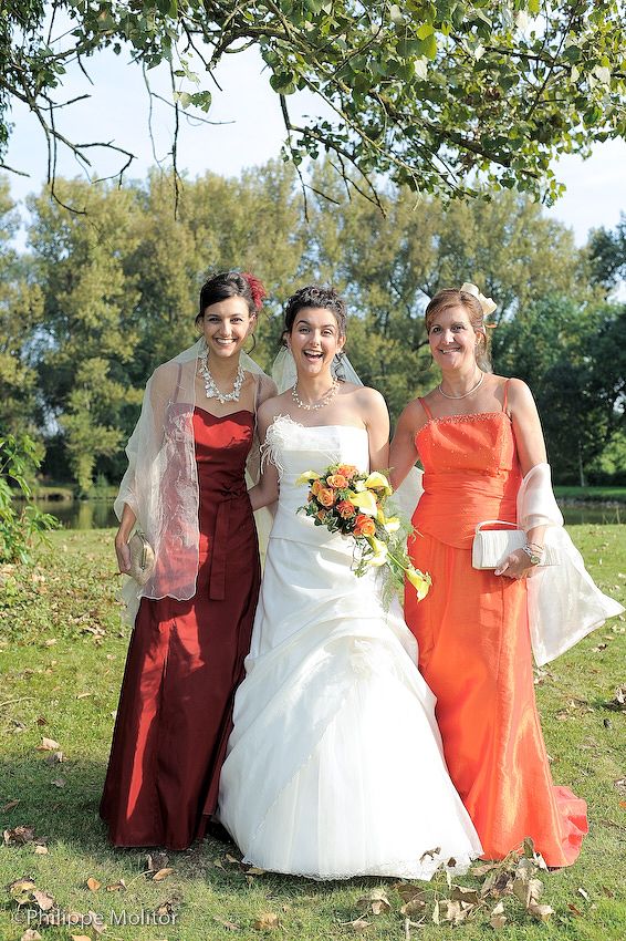 Bride in white gown with two bridesmaids in red and orange dresses, holding bouquet outdoors at wedding.