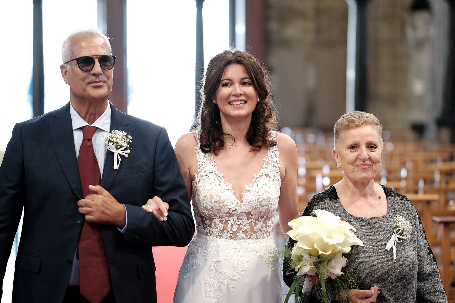 Bride in white lace gown walks down church aisle flanked by older man in suit and elderly woman holding roses.