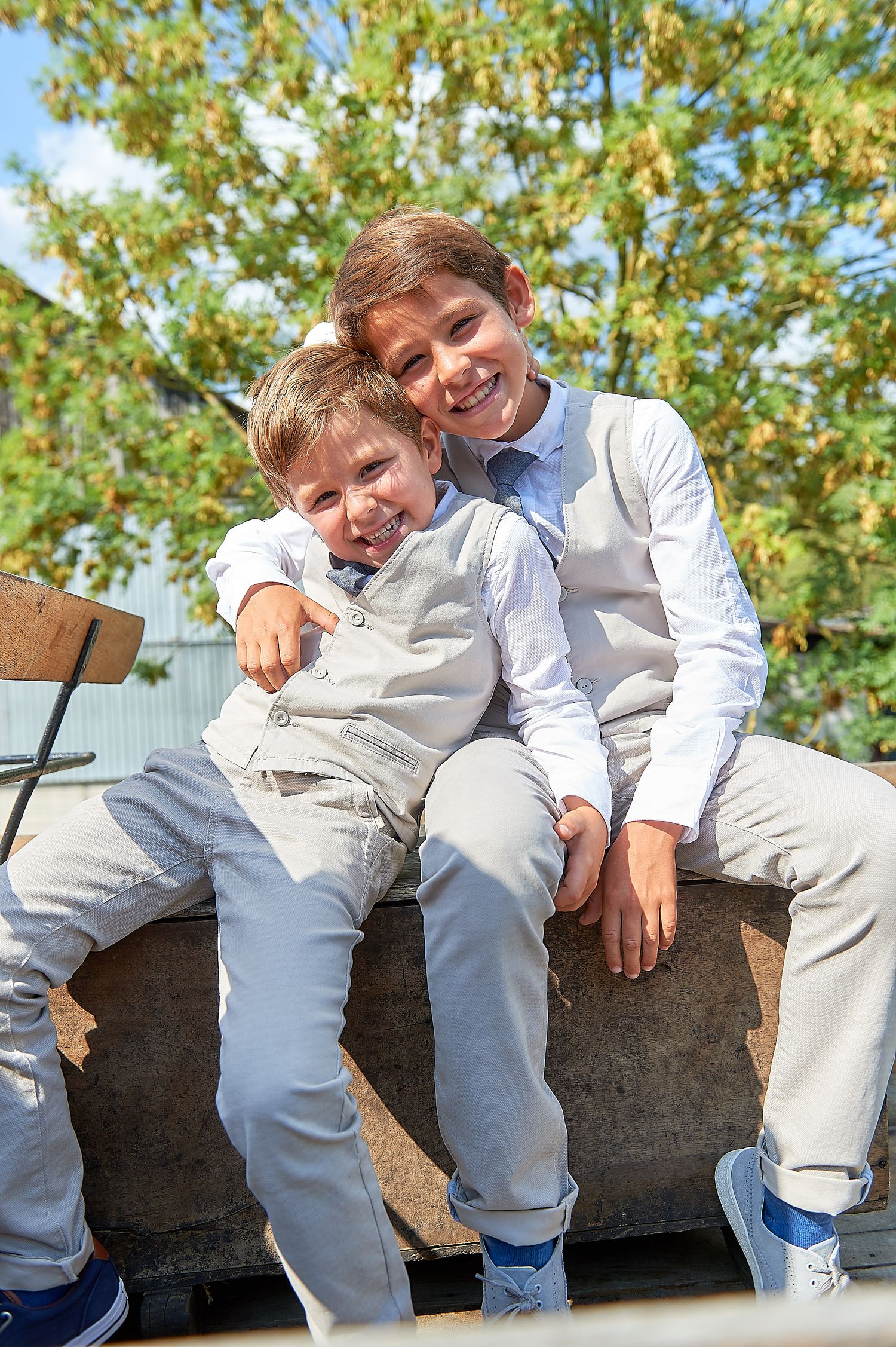 Two smiling boys in matching white outfits sitting together outdoors, with green trees in the background.