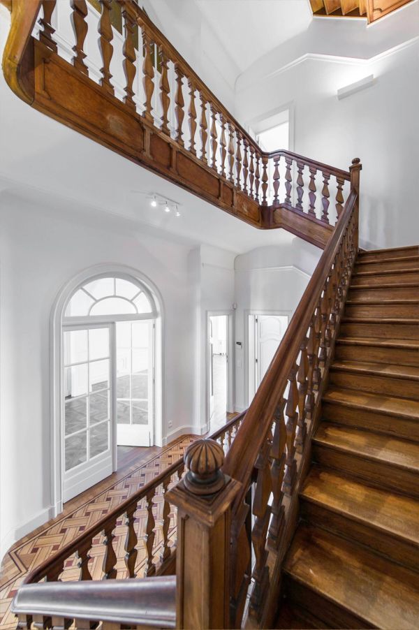 Grand wooden staircase with ornate balusters and arched window in bright white interior.