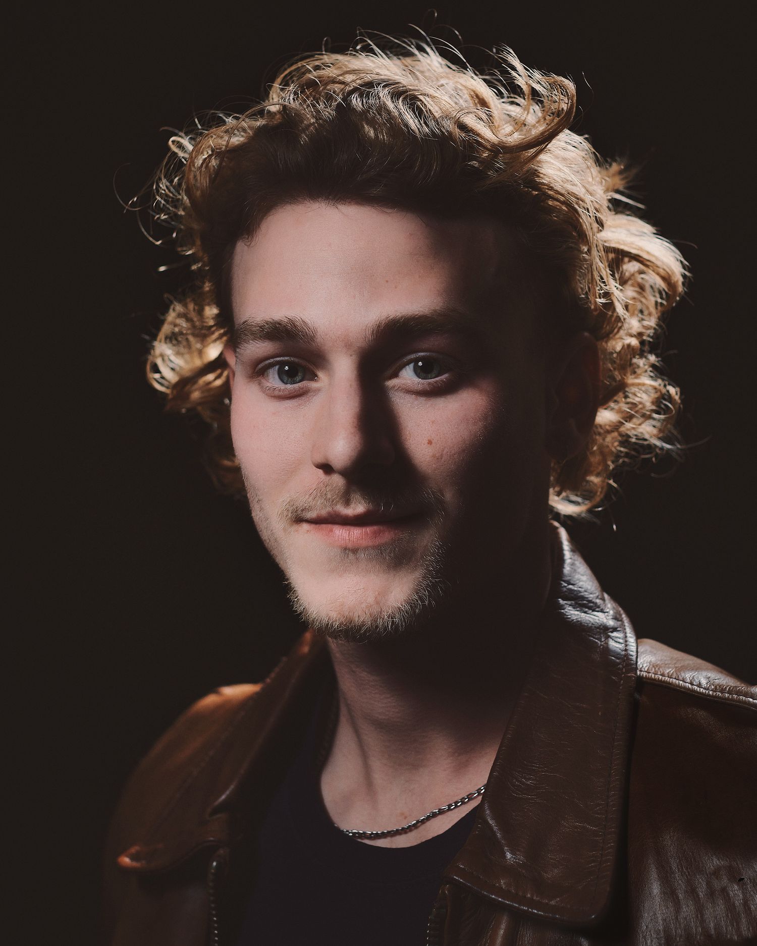 Portrait of someone wearing leather jacket with curly hair against dark backdrop.