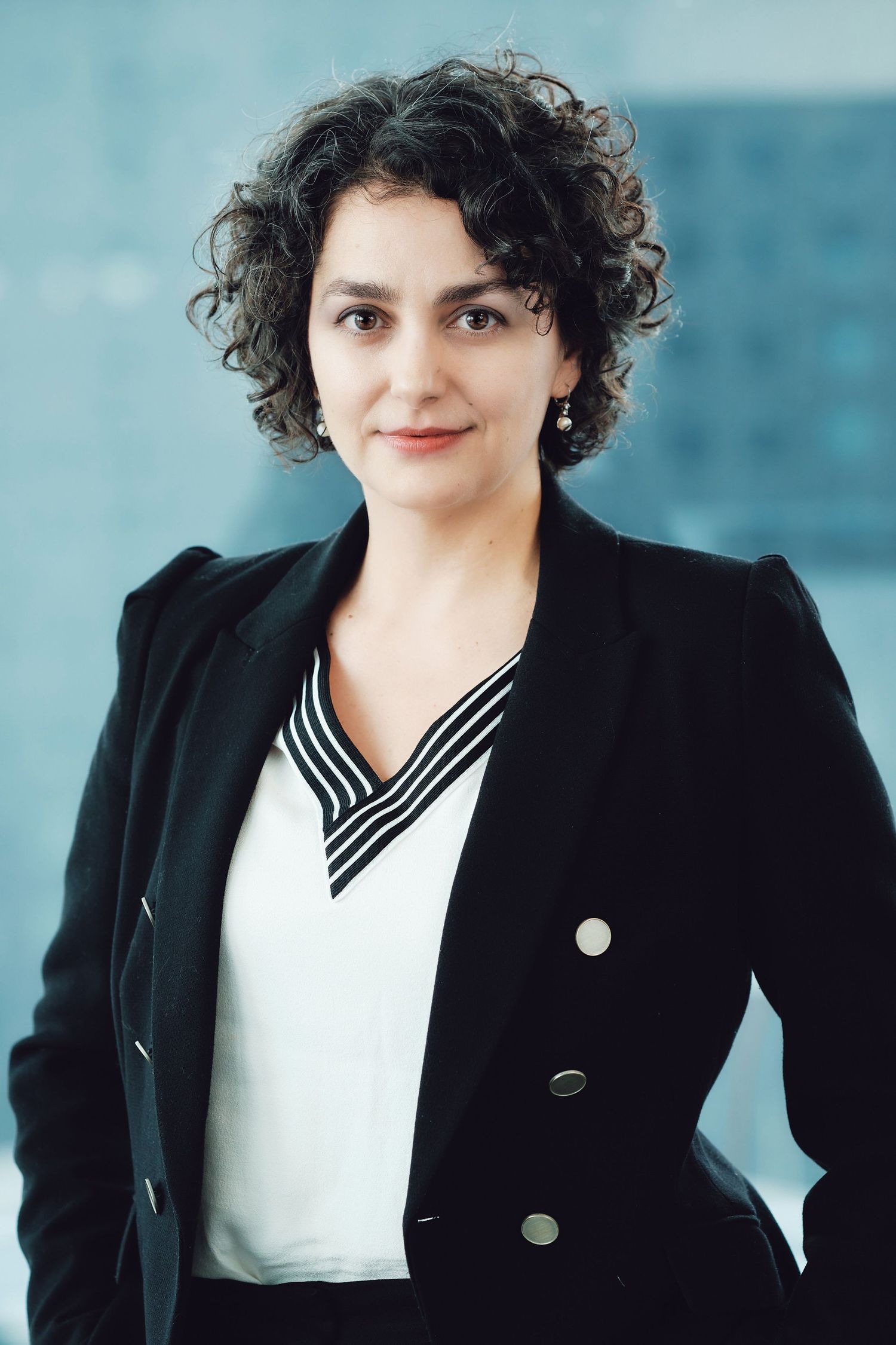 Corporate headshot of a professional wearing a black blazer and striped collar blouse against blue backdrop.