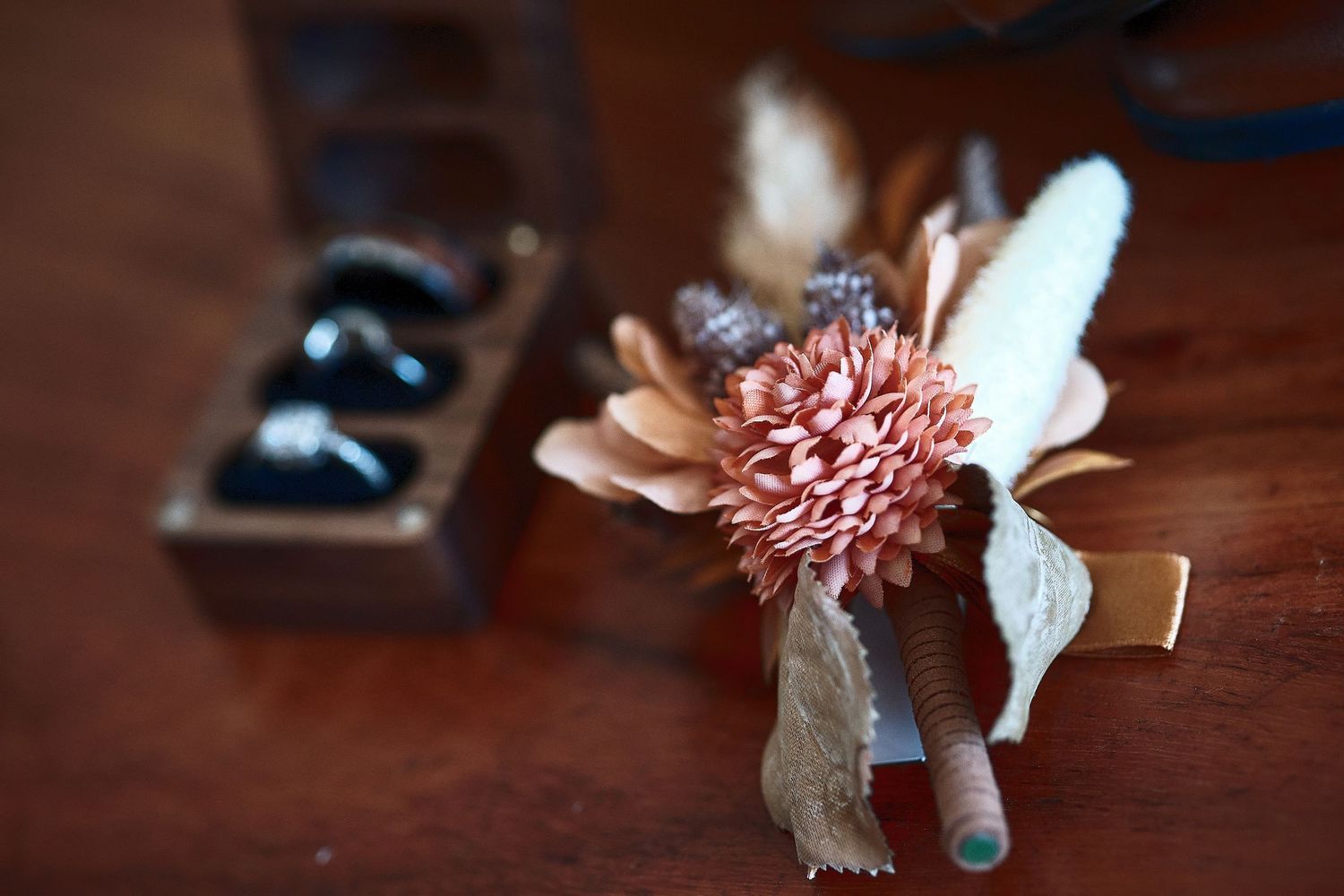 Rustic boutonniere with pink dahlia, feather, and burlap ribbon on wooden table near ring box.