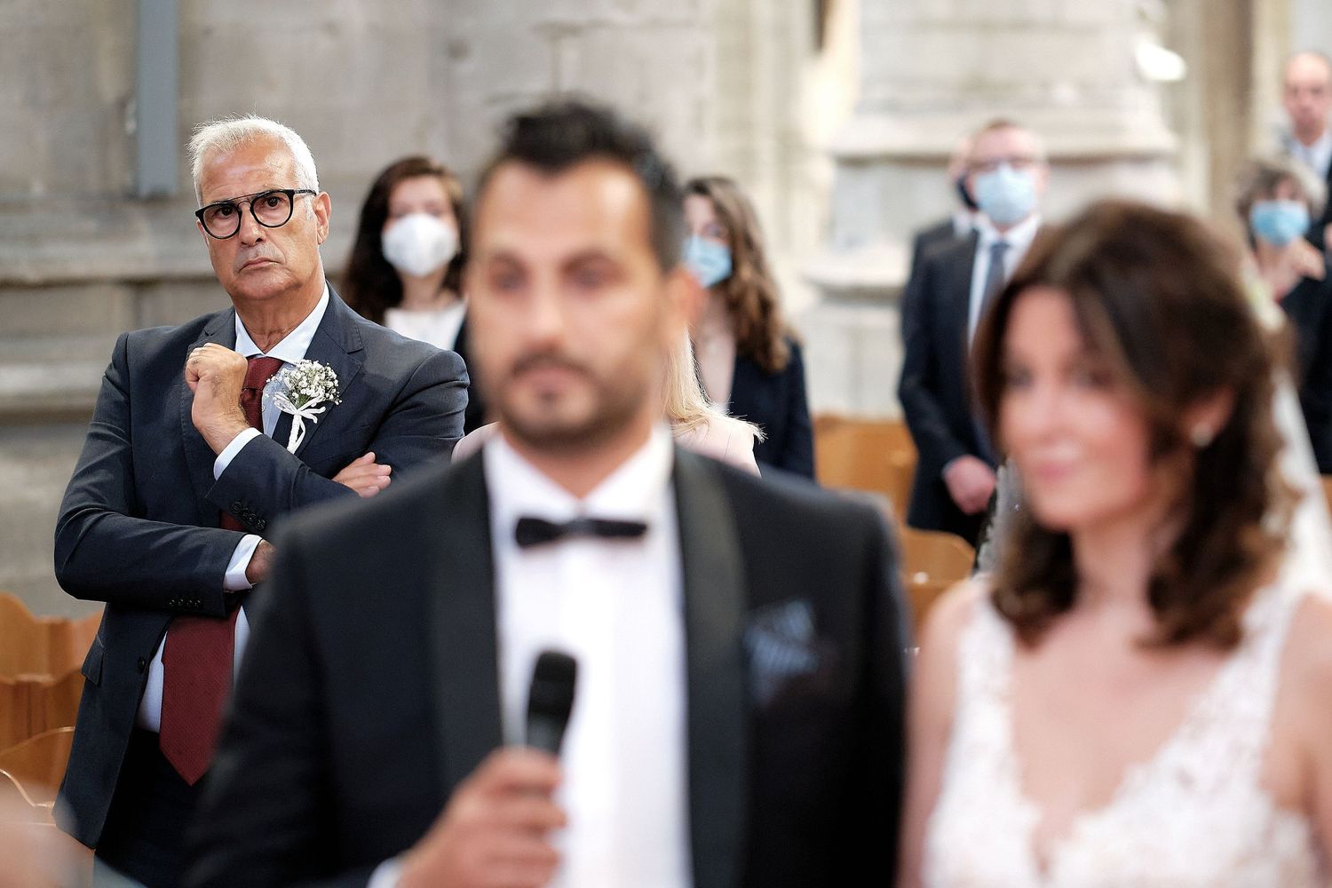 Wedding ceremony with guests wearing masks, groom speaking at microphone while father-in-law watches with crossed arms.