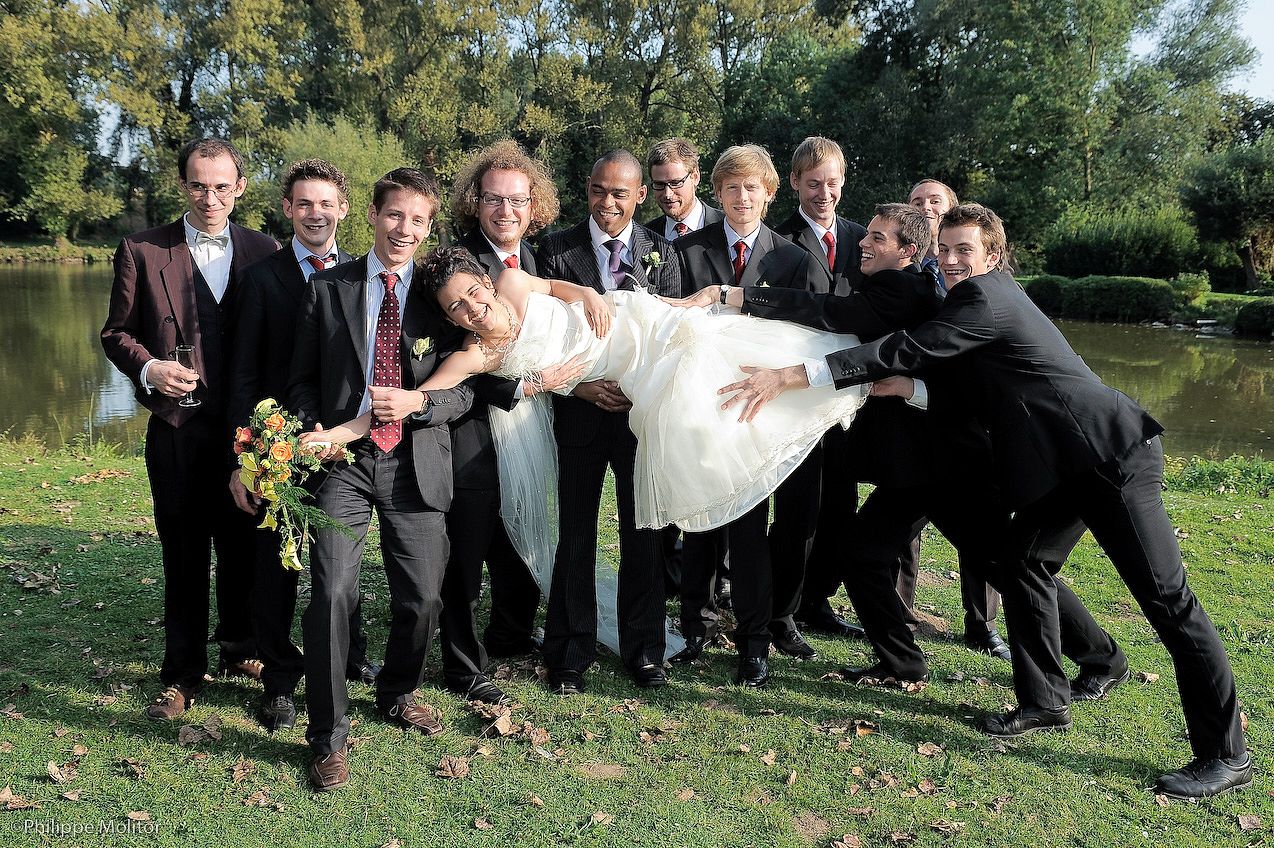 Groomsmen playfully lifting bride in white dress outdoors near pond at wedding celebration.