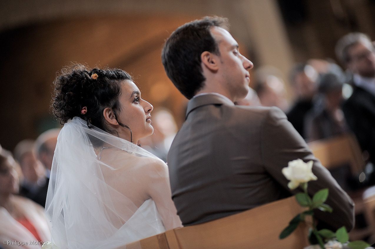 Bride in white veil gazes forward during church wedding ceremony while groom sits beside her in a brown suit.