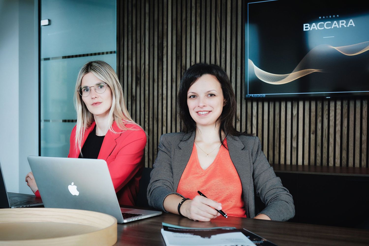 Professional in a red blazer and dark top sits at a desk during a business meeting.