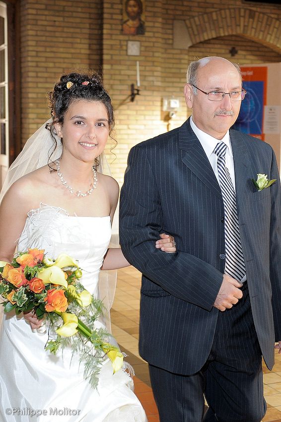 Bride in white gown holding orange bouquet walks arm-in-arm with older man in suit at church wedding.