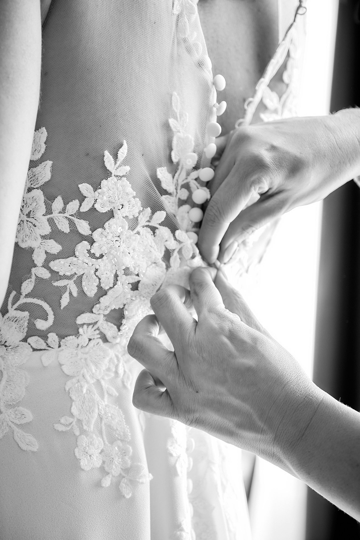Black and white photo of hands fastening buttons on a lace wedding dress back.