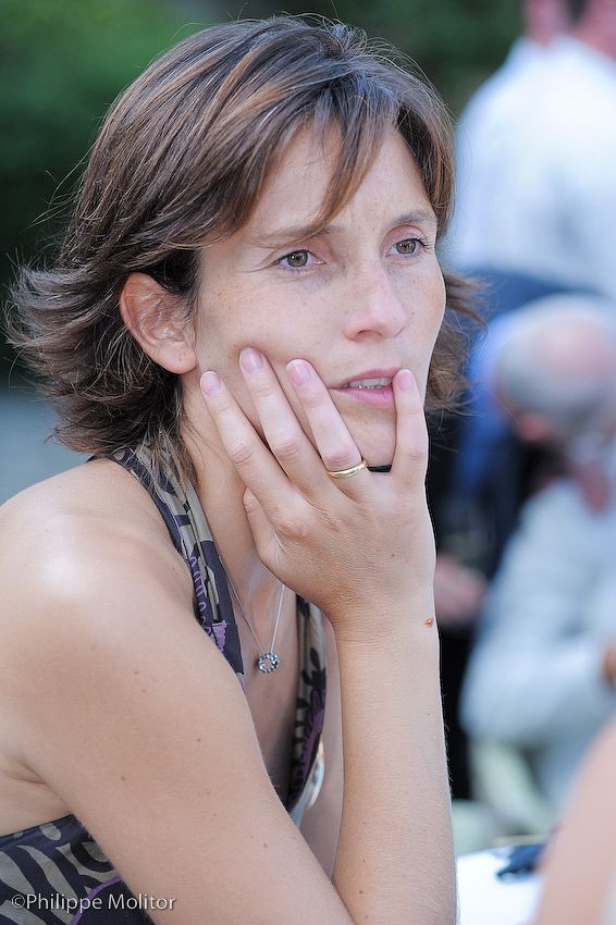 A thoughtful woman resting her chin on her hand, wearing a necklace and ring, with a blurred outdoor background.