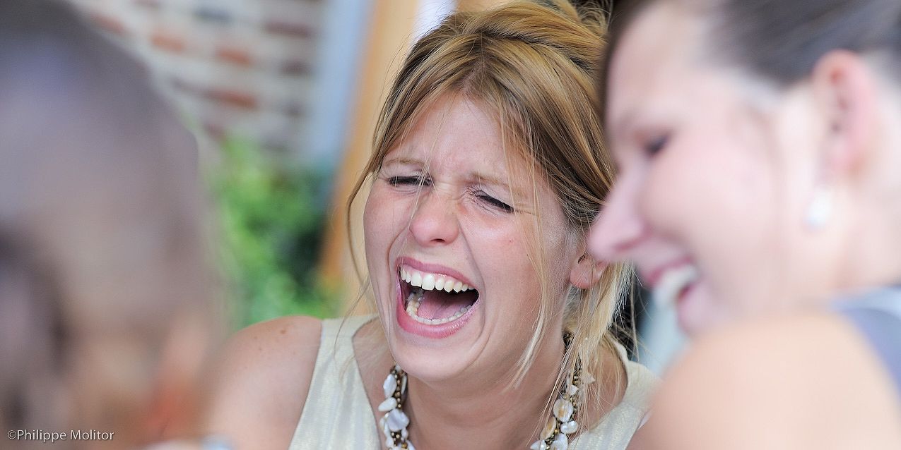 A woman laughing joyfully at a social gathering, wearing a white top and statement necklace.