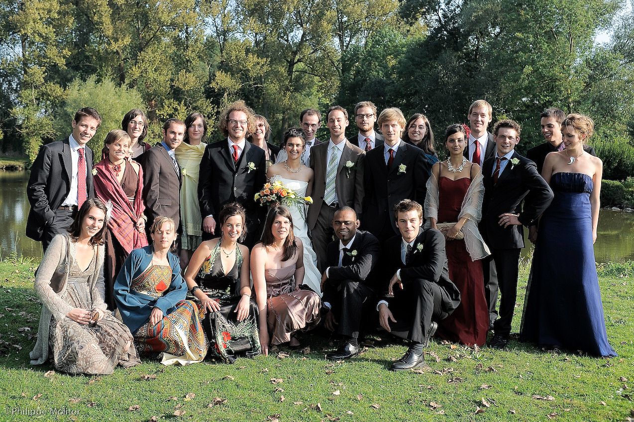 Large wedding party group photo outdoors near a pond, featuring bride, groom, and guests in formal attire.