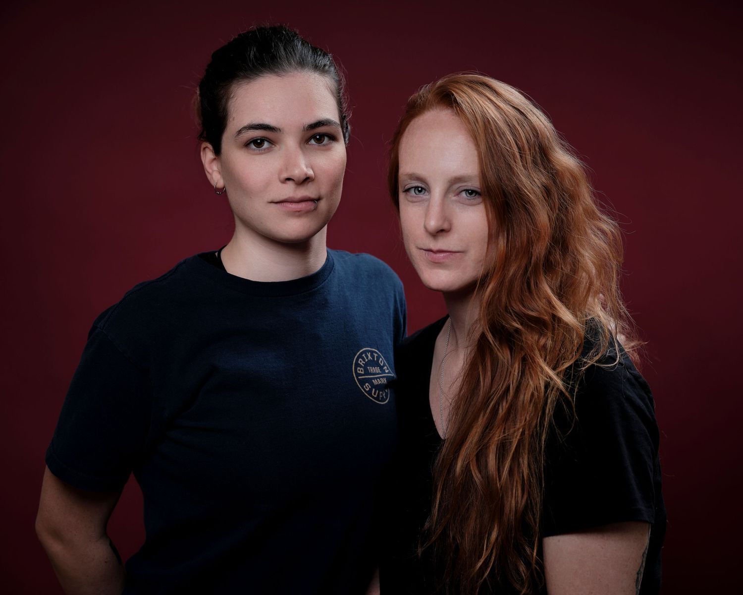 Two women posing together against a dark red background, one with dark hair and one with long red hair.