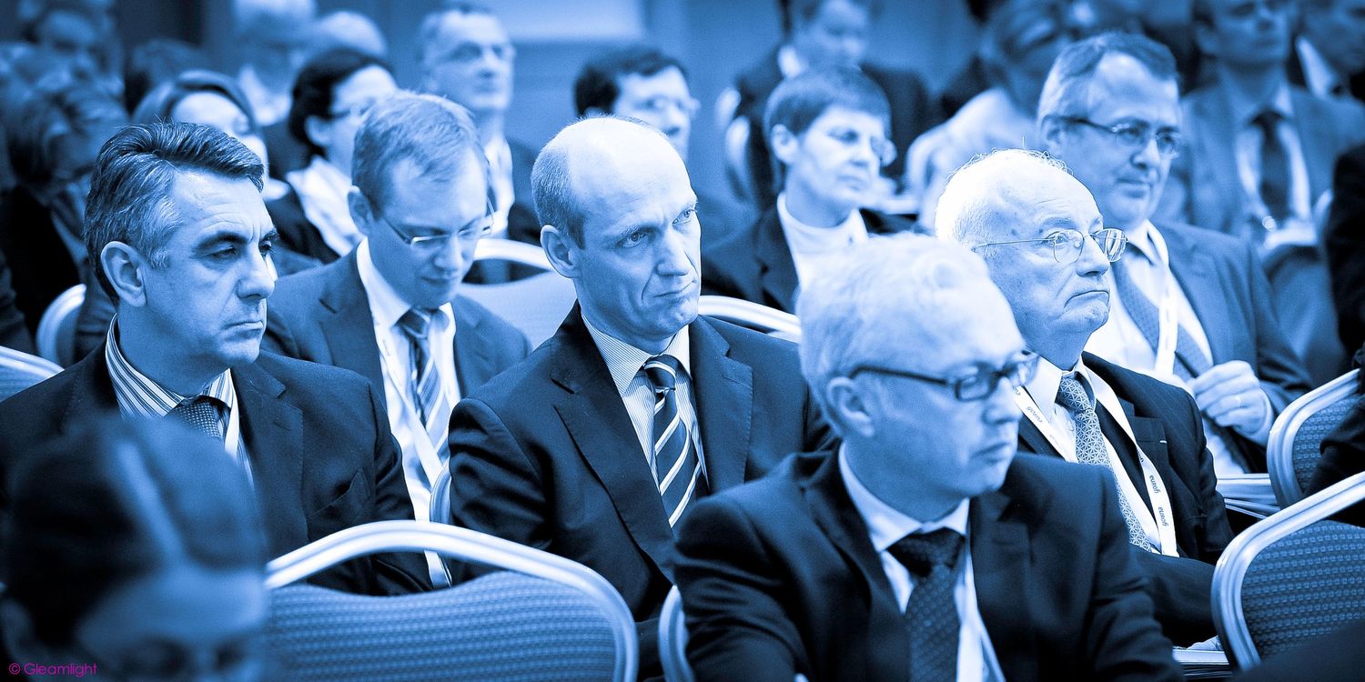 Business conference attendees in formal attire shown in blue-tinted panoramic view.