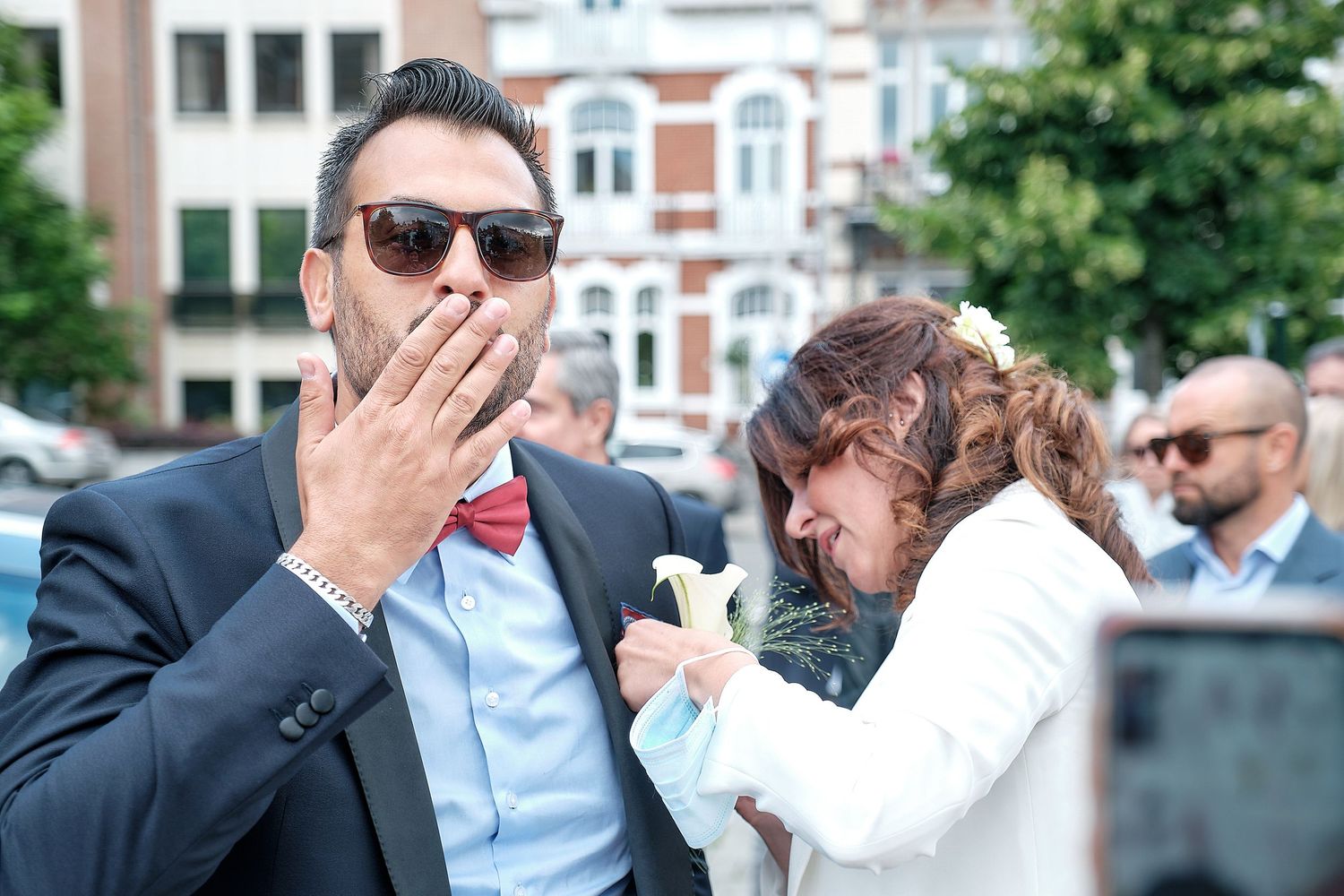 A man in a suit with a red bow tie covers his mouth while a woman in white stands beside him outdoors.
