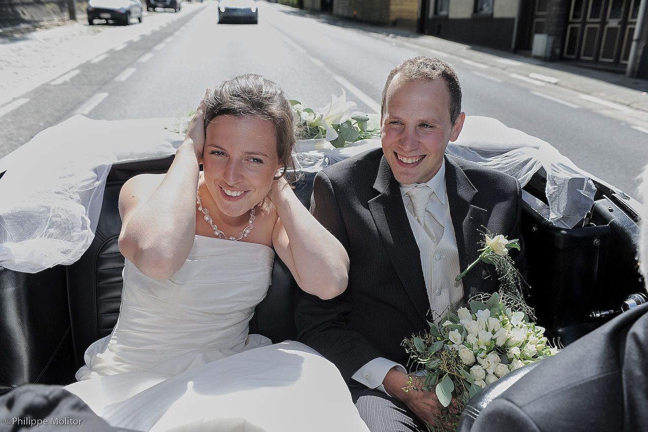 Happy newlyweds ride in a convertible car, bride in white dress holding bouquet, groom in suit on their wedding day.