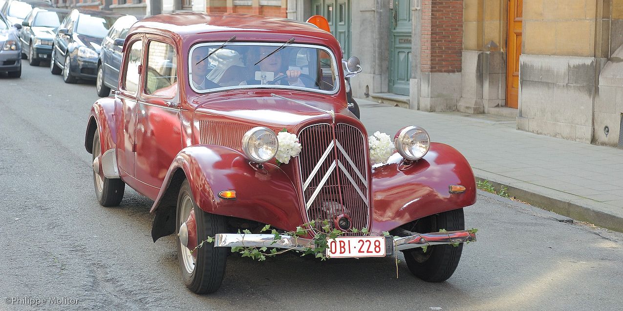 Vintage red Citroën Traction Avant decorated with flowers drives down a cobblestone street in Belgium.