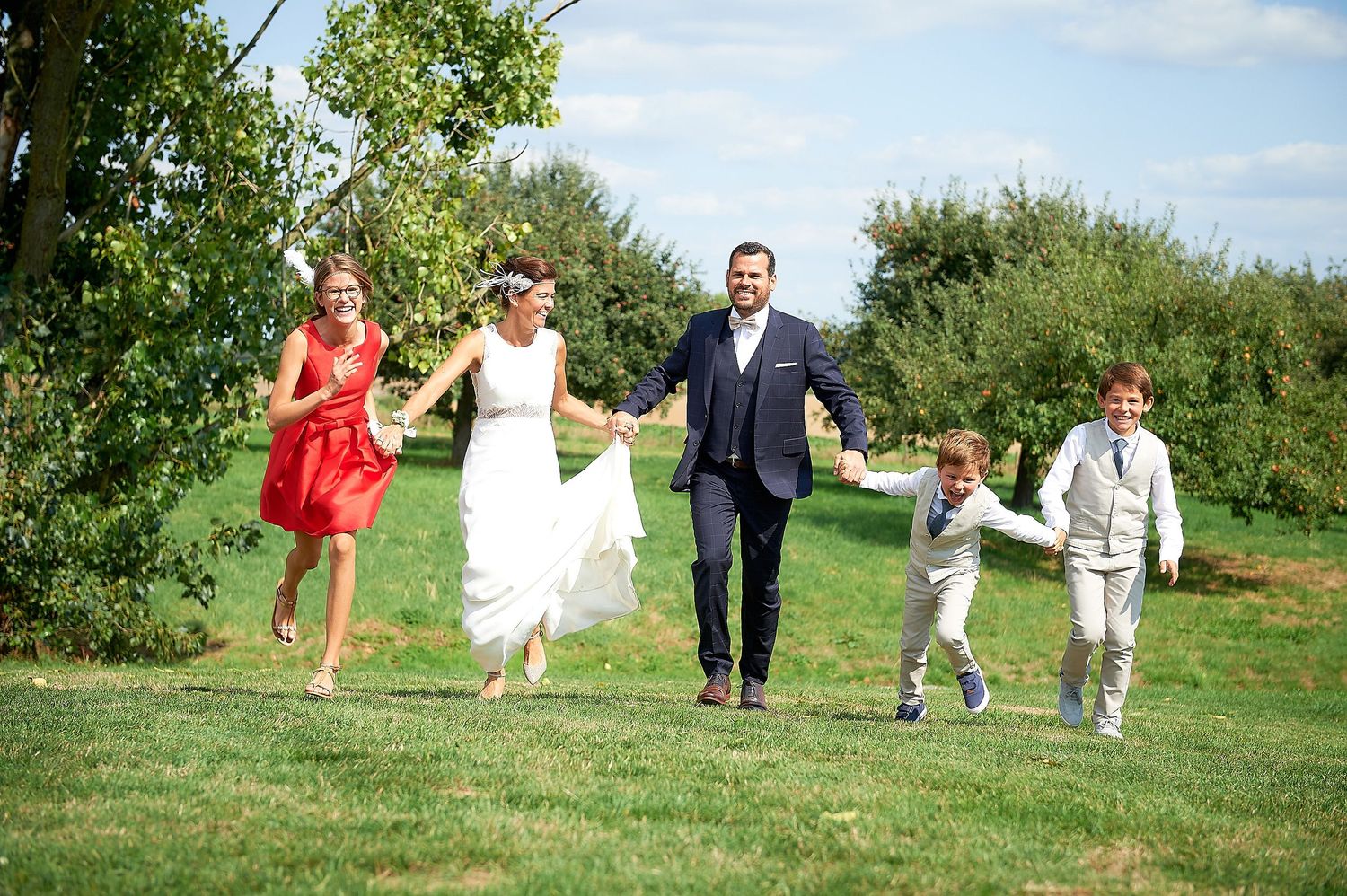 A joyful wedding party runs hand-in-hand across a green lawn, featuring a bride, groom, and three children.