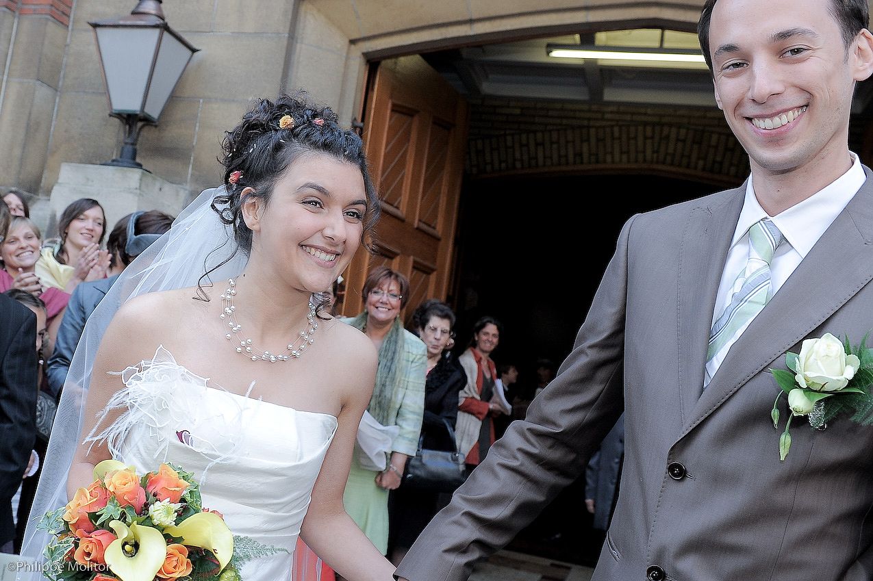 Happy newlyweds exit church after wedding ceremony, bride holding colorful bouquet, groom in gray suit with boutonniere.