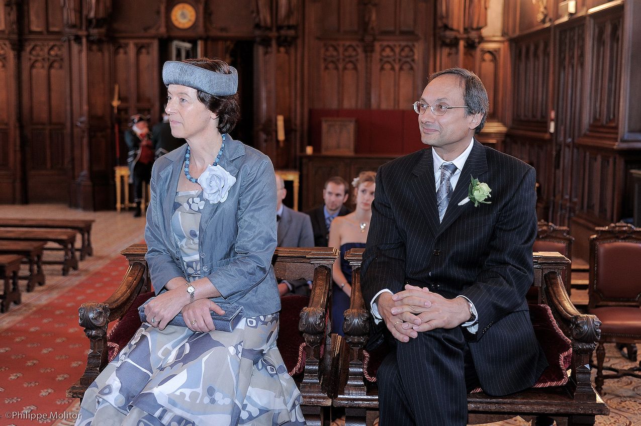 Elegant wedding guests seated in ornate church pews, woman in blue dress with hat beside man in dark suit.