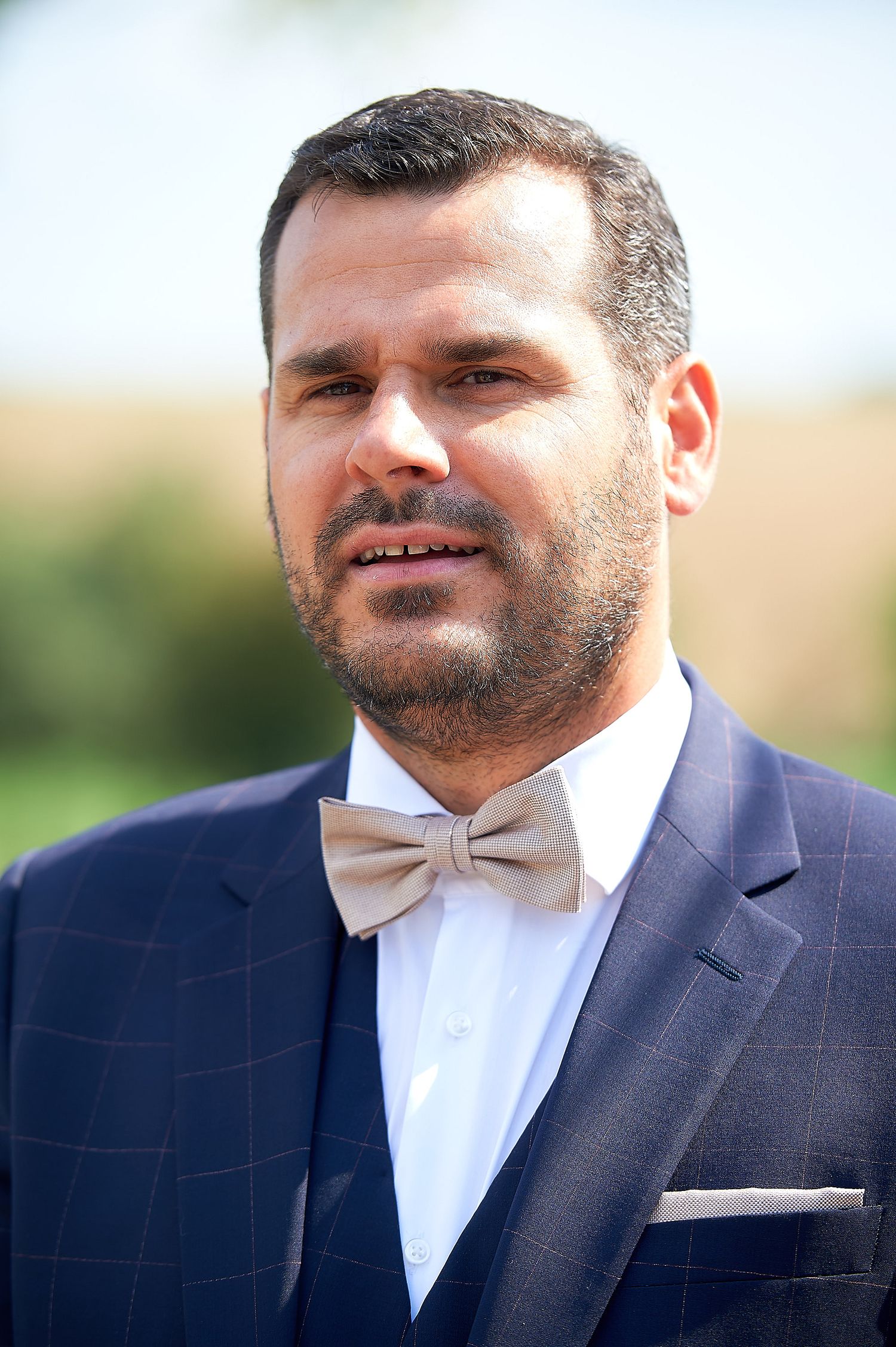 Man in navy blue plaid suit with beige bow tie and beard posing outdoors at a formal event.