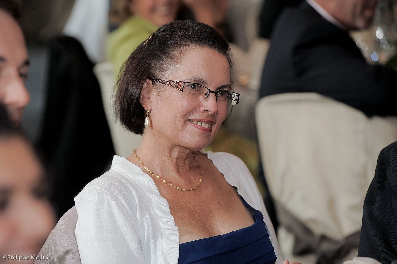 Woman with glasses and short brown hair wearing a blue dress and white shawl with a gold necklace at an event.