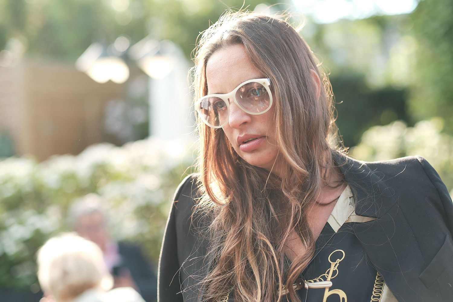 Stylish woman with long brown hair wearing oversized white sunglasses and black outfit outdoors.