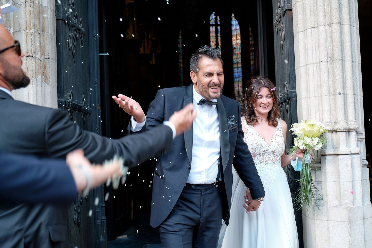 Joyful newlyweds exit church doors as guest throws confetti during outdoor wedding celebration.