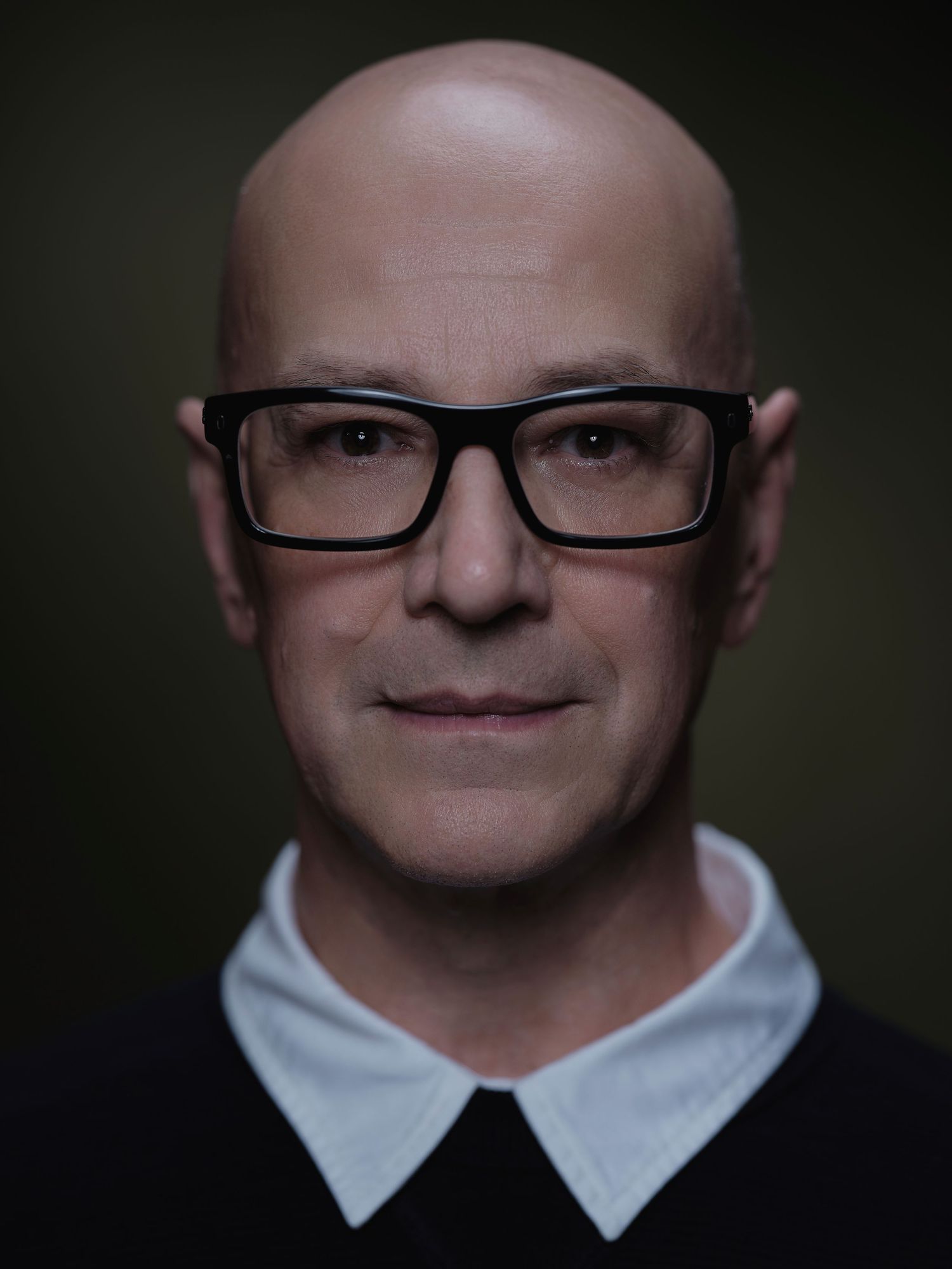Bald man with black-framed glasses wearing a black top with white collar, photographed against dark background.