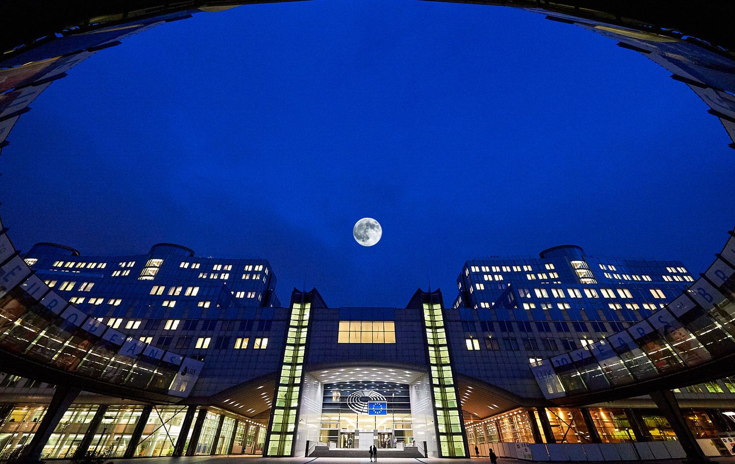 Modern office building exterior at night with illuminated windows and full moon against deep blue sky.