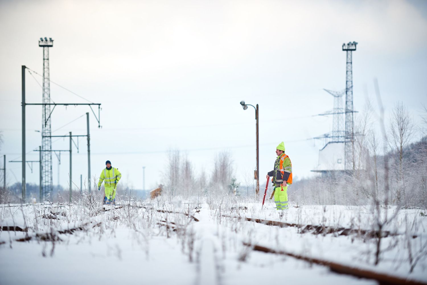 Maintenance workers in safety vests inspect snow-covered railway tracks with power lines in background.