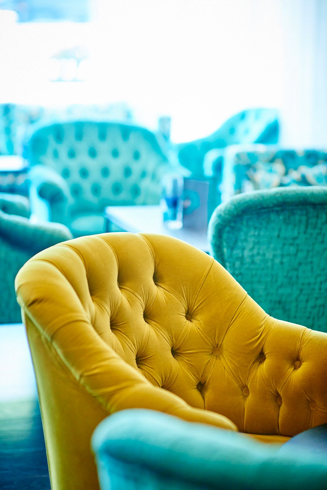 Close-up of yellow tufted velvet chair with turquoise furniture in background.
