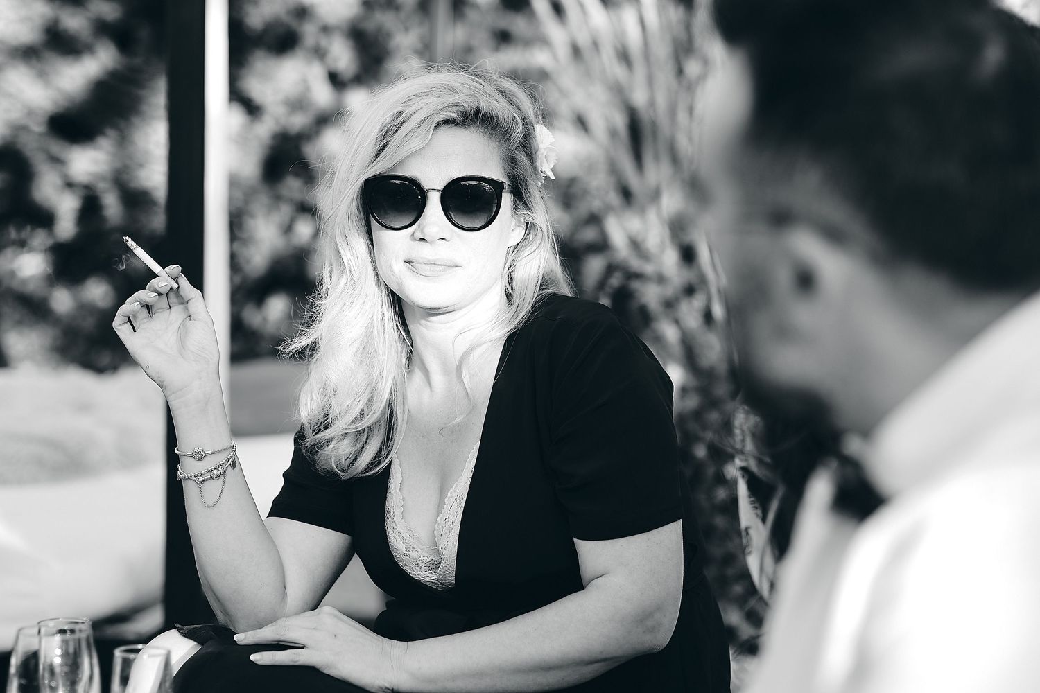Black and white photo of a blonde woman in sunglasses holding a cigar at an outdoor cafe.