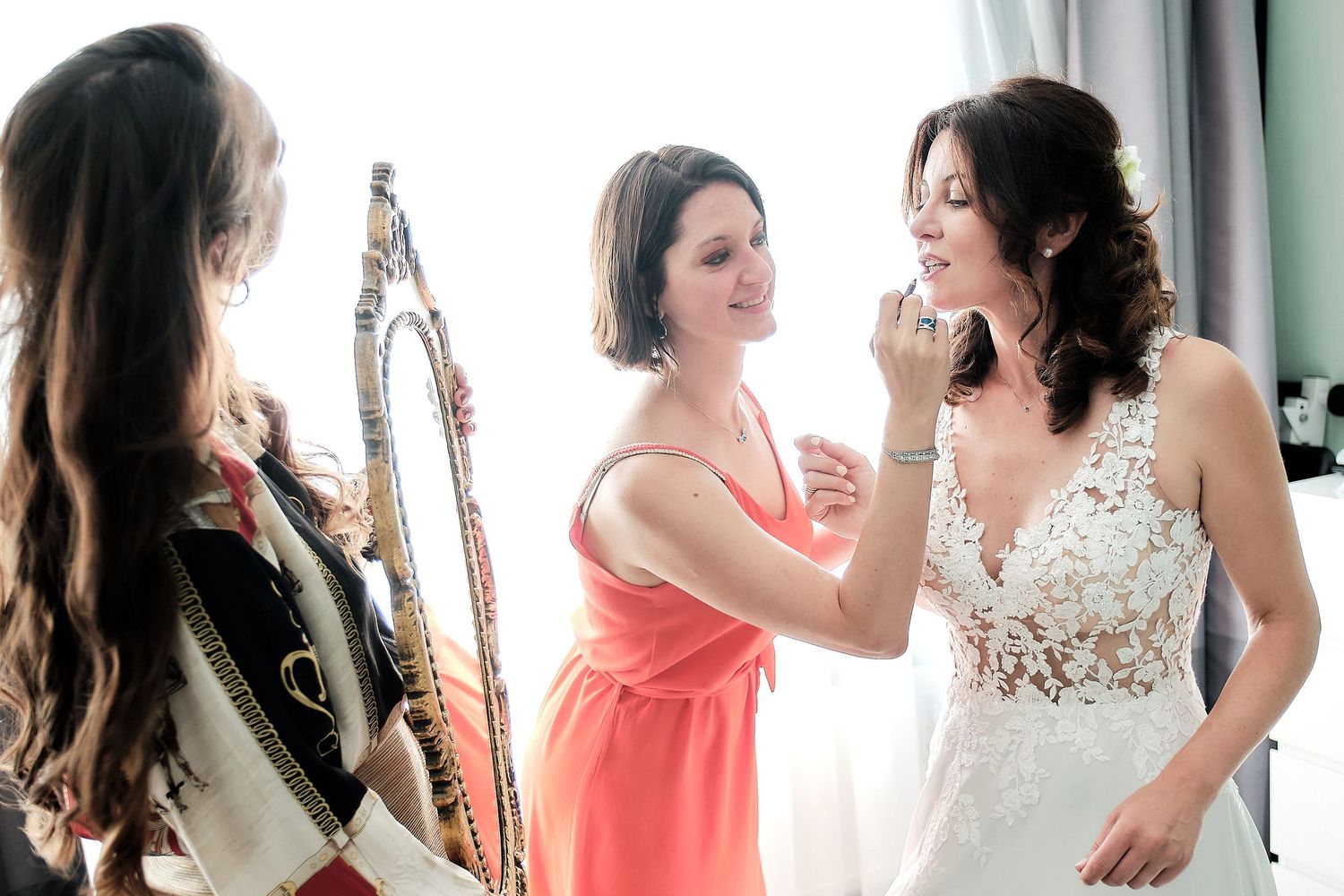 Bridesmaid in coral dress helping bride in lace wedding gown prepare, with another woman watching near a mirror.