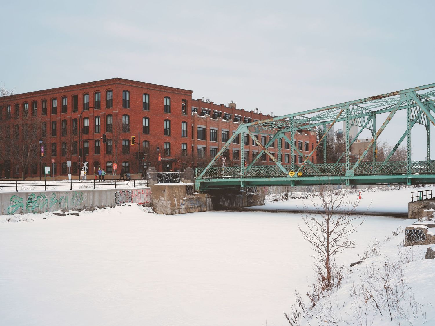 Snow-covered riverbank with red brick buildings and green metal bridge spanning across frozen waters.