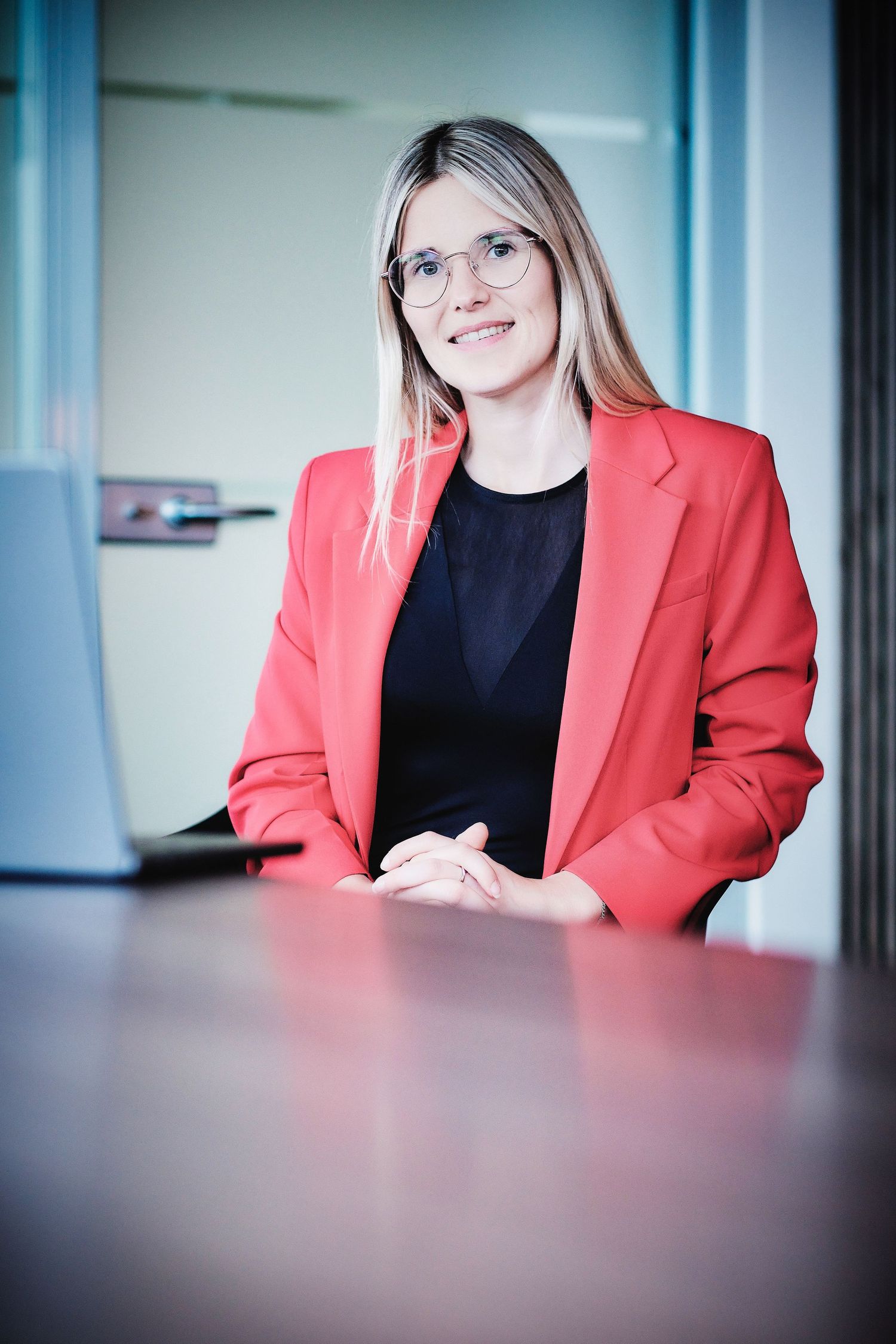 Person in coral blazer and black top seated at modern office desk with blurred foreground.
