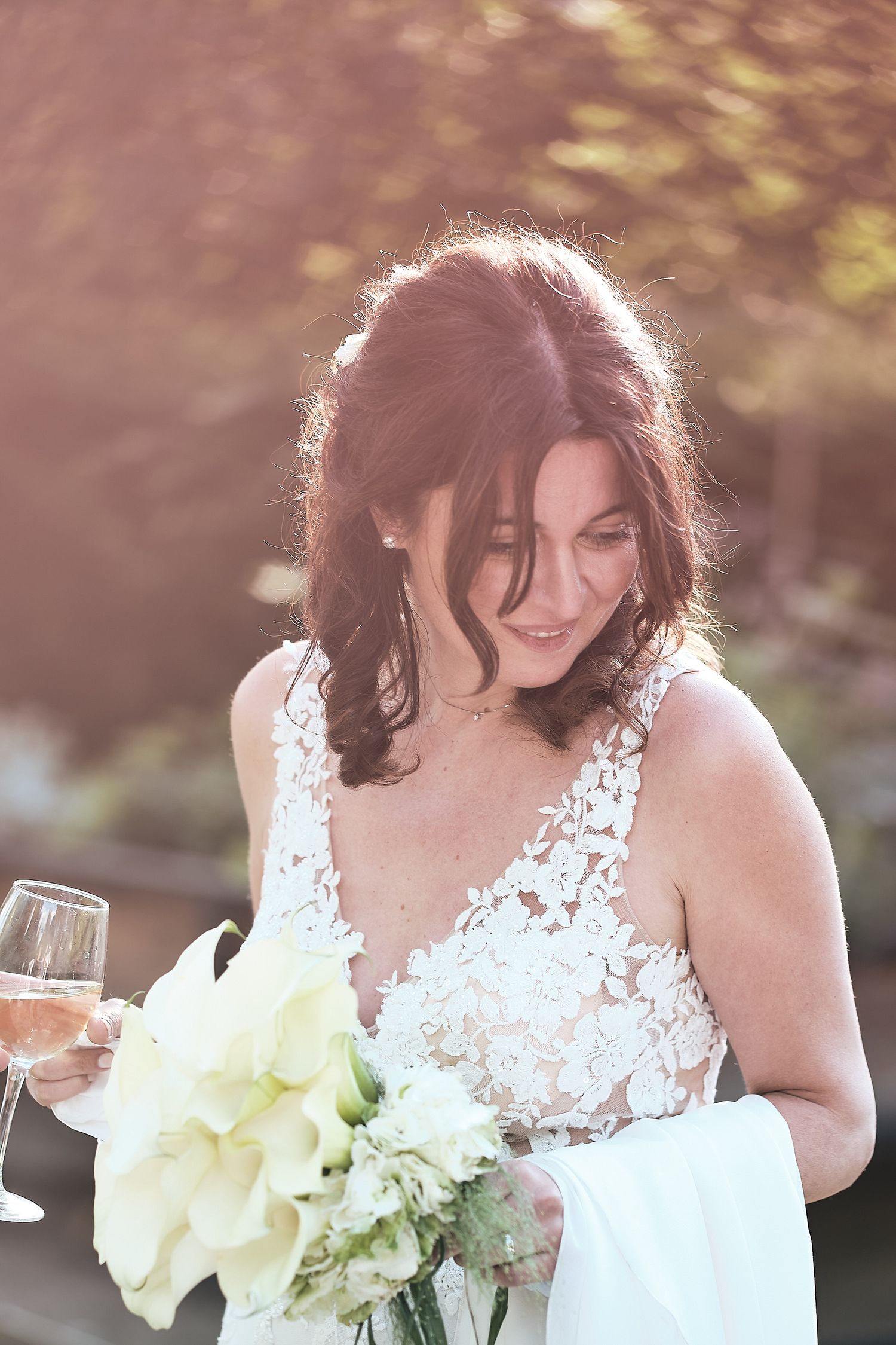 Smiling bride in lace wedding gown holding white flower bouquet and champagne glass outdoors in sunlight.