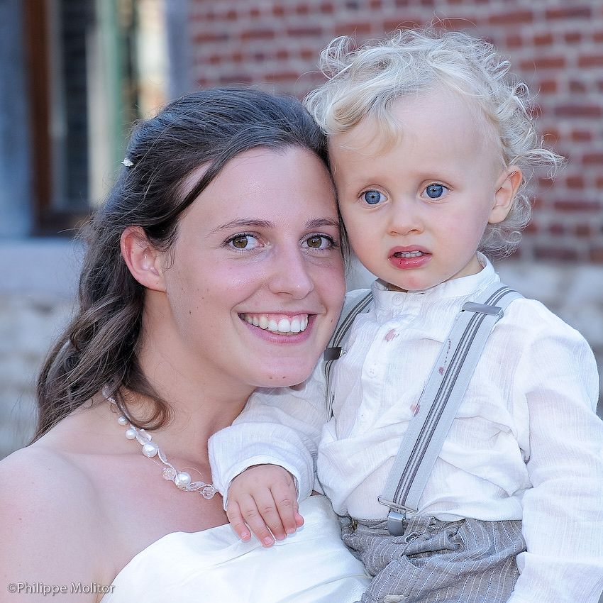 A smiling bride in white dress holding a curly-haired toddler boy with blue eyes wearing suspenders at a wedding.