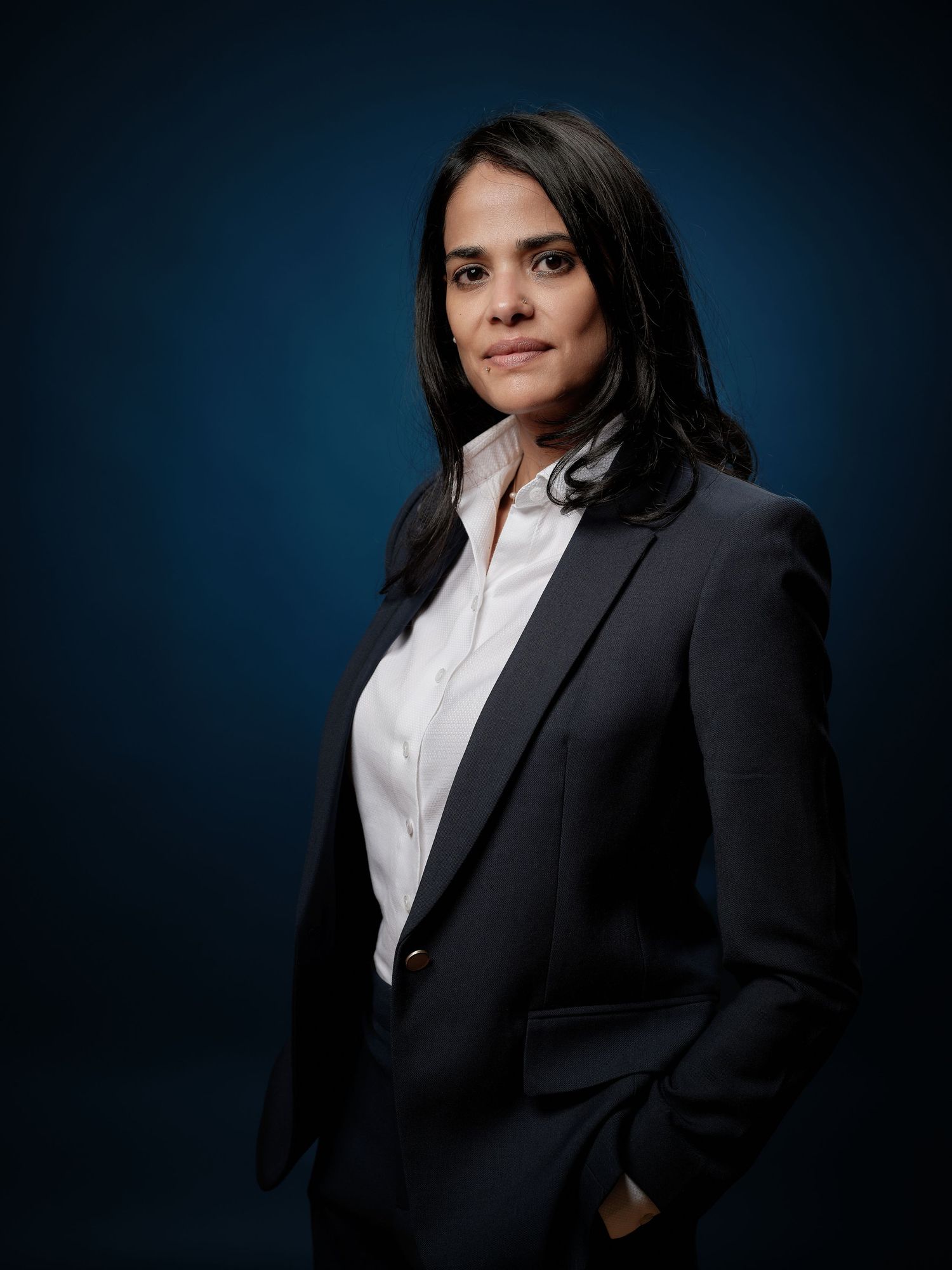 Professional woman in dark blazer and white shirt poses confidently against a dark blue background.