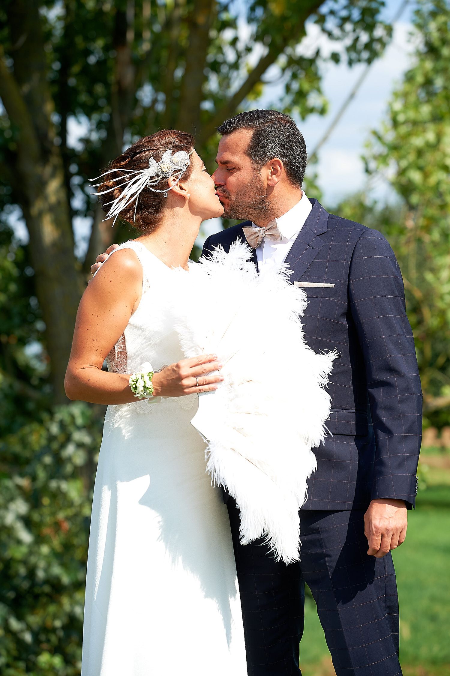 Bride and groom sharing a romantic kiss outdoors, bride holding white feather fan and wearing feather hair accessory.