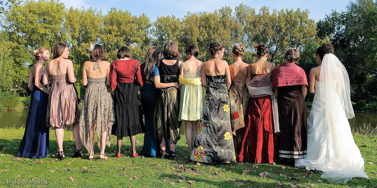 Bride and bridesmaids in colorful dresses standing with their backs to camera in a garden near a pond.