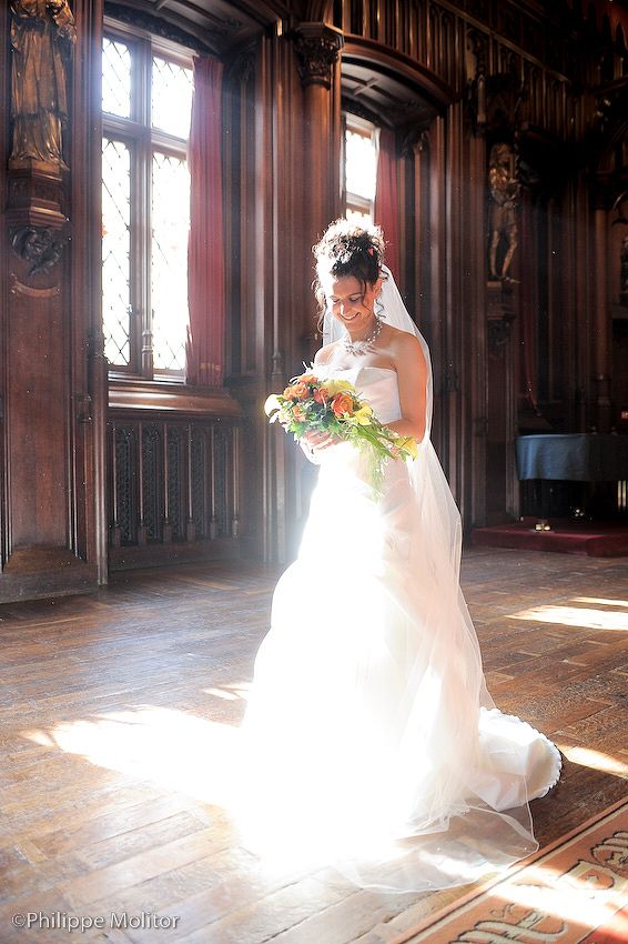 Elegant bride in white ball gown holds colorful bouquet in sunlit ornate wood-paneled hall with stained glass windows.