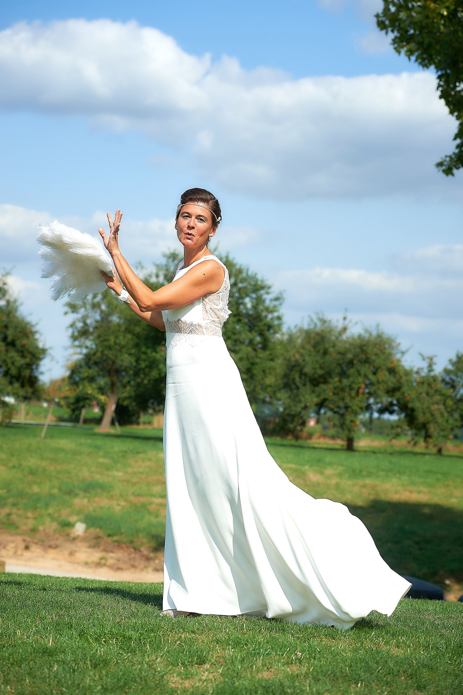 Bride in white gown tossing bouquet outdoors on green lawn with blue sky and trees in background.
