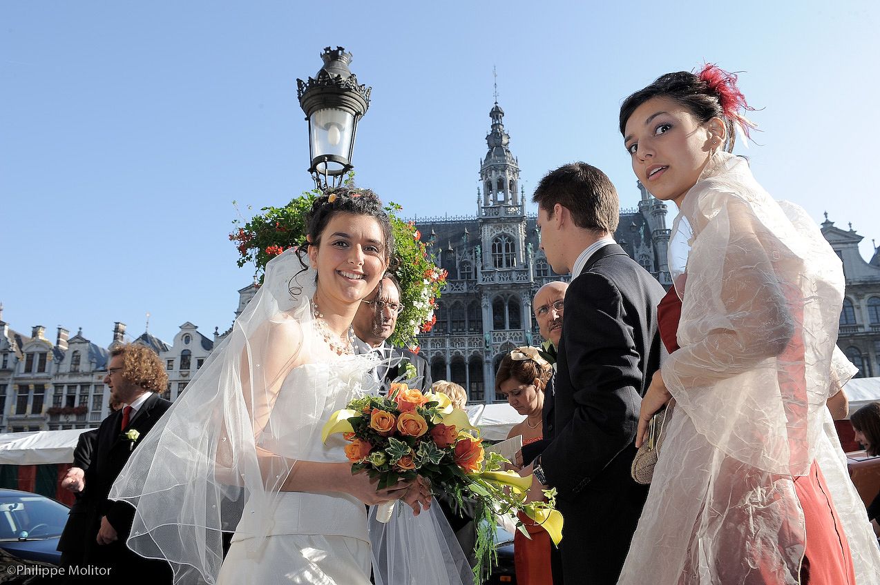 Bride in white veil holding colorful bouquet stands with guests in elegant attire at Grand Place, Brussels.