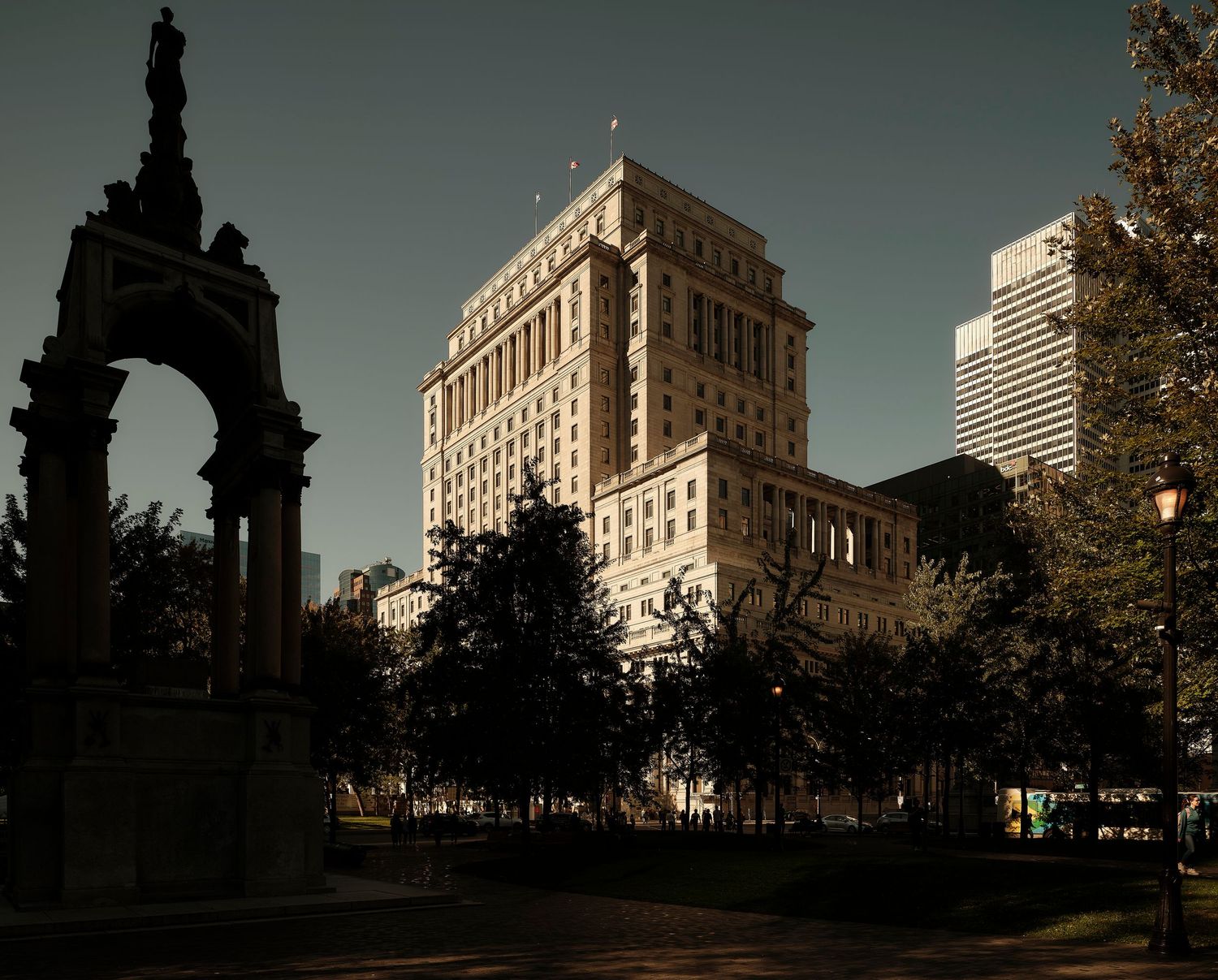 Historic monument and grand building at dusk in a city park, surrounded by trees with a dramatic dark sky.