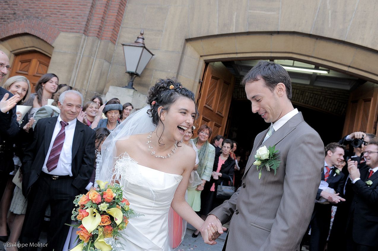 Happy bride and groom holding hands leaving church after wedding ceremony, guests celebrating with colorful bouquet.