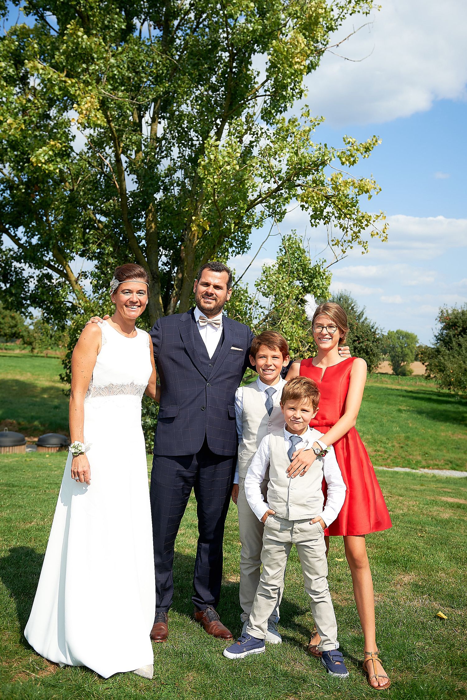 Family posing outdoors at a wedding, bride in white gown, groom in navy suit, two boys and a woman in red dress.