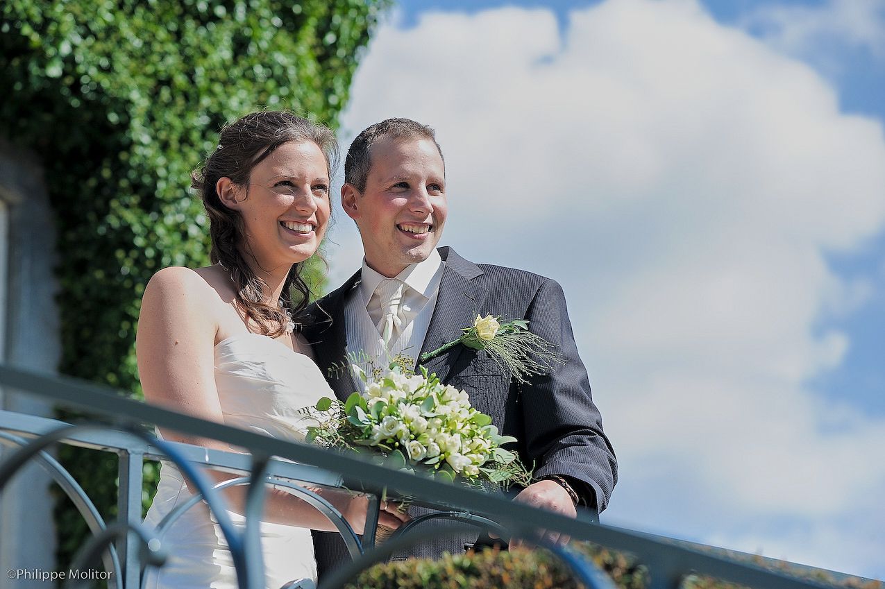 Happy newlywed couple smiling on a balcony, bride holding white bouquet, groom in gray suit with boutonniere.