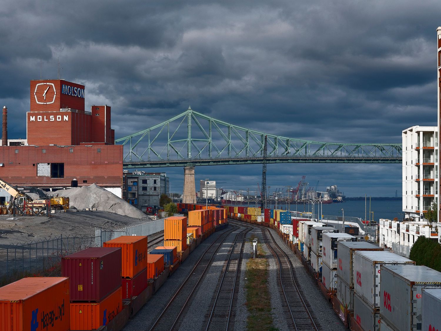 Industrial port scene with shipping containers and railroad tracks beneath stormy skies and Montreal's Jacques Cartier Bridge.