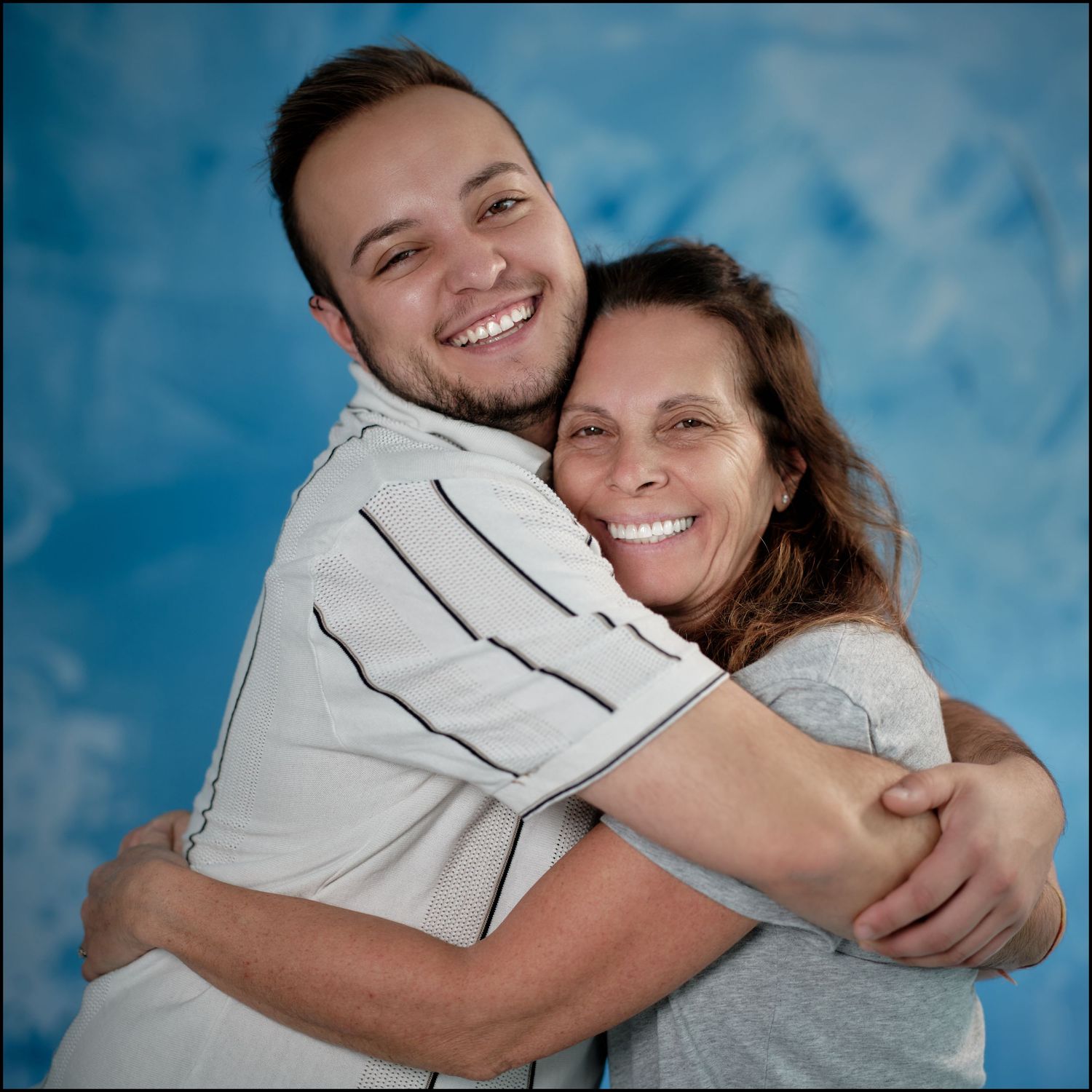A young man and an older woman share a warm hug, smiling joyfully against a blue background.
